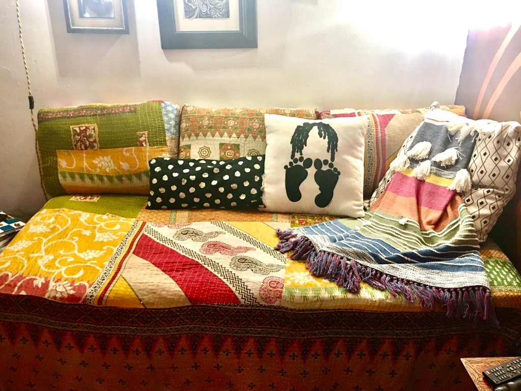 Two images. One of the twin bed covered with how pillows and the custom cover with red and yellow bands of color and a paisley design. The other image shows the Ottoman with the custom cover on it featuring green and fold colors and geometric design elements.