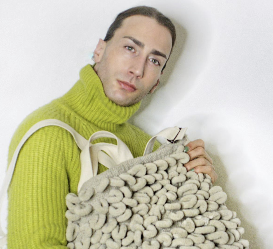 Peer Cox Headshot wearing a bright green turtleneck sweater with a bag made of thick yarns.